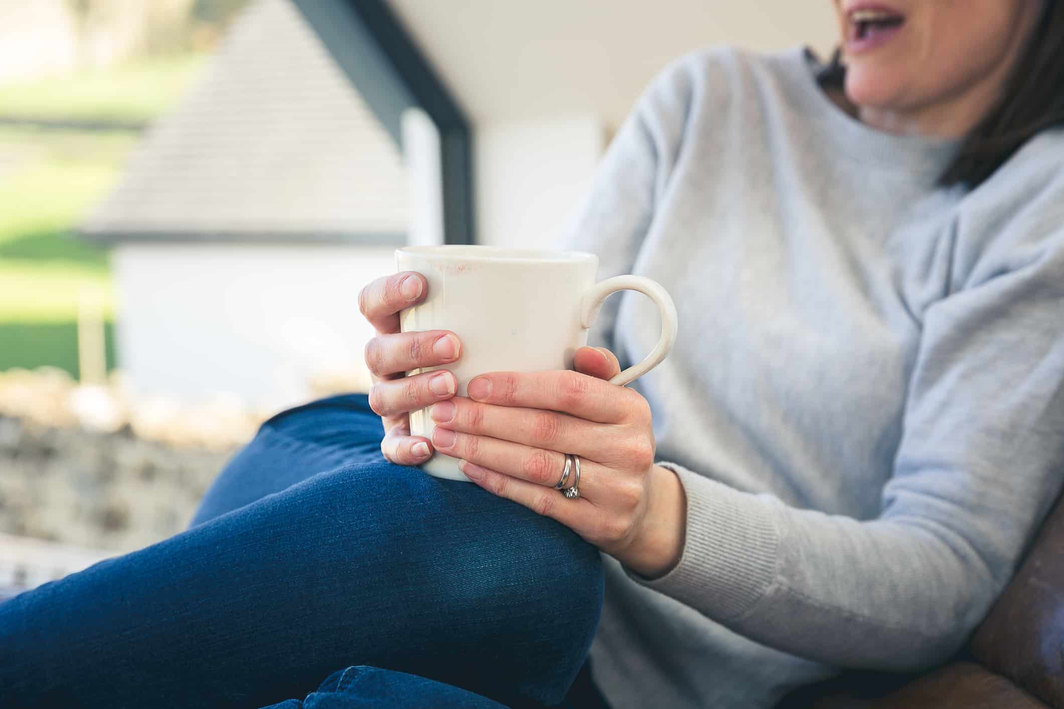 rewind-trauma-therapy-online-hypnotherapy white woman, wearing blue jeans, grey jumper holding a white cup upper half of face out of shot to demonstrate the absence of trauma talk during trauma rewind therapy