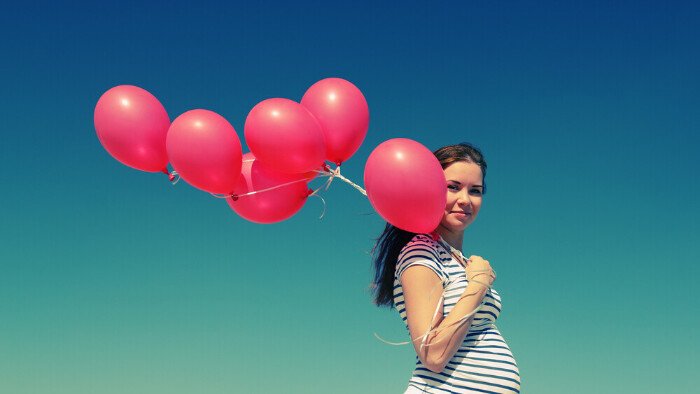 Hypnobirthing-avoid-negativity white pregnant woman holding bright pink balloons and smiling positively to demonstrate the positive emotions of feeling confident in pregnancy and birth