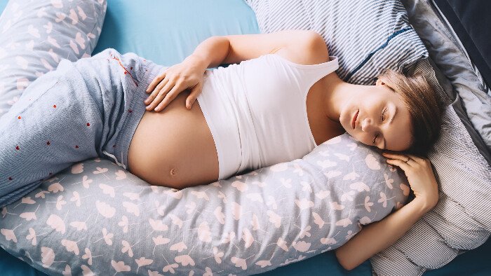 Hypnobirthing-comfort-measures An example of a calm birth setting and hypnobirthing comfort measures a white pregnant woman is lying on her side in bed using a pregnancy pillow for support and resting deeply