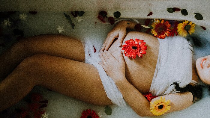 Hypnobirthing-fear-release-techniques white pregnant woman with long brown hair modestly covered with white cloth and relaxing in a milk bath with flowers demonstrating the relaxed confidence that comes from the hypnobirthing practice of gentle fear release