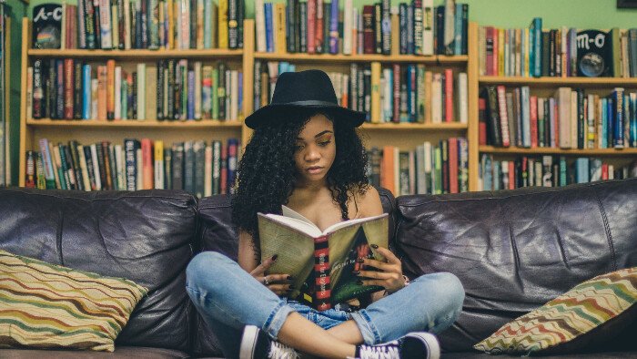 Informed-birth-knowledge black woman in blue jeans and a wide brimmed black hat sits on a leather sofa reading a birth book in a library to improve her birth knowledge and reduce her fear of pain