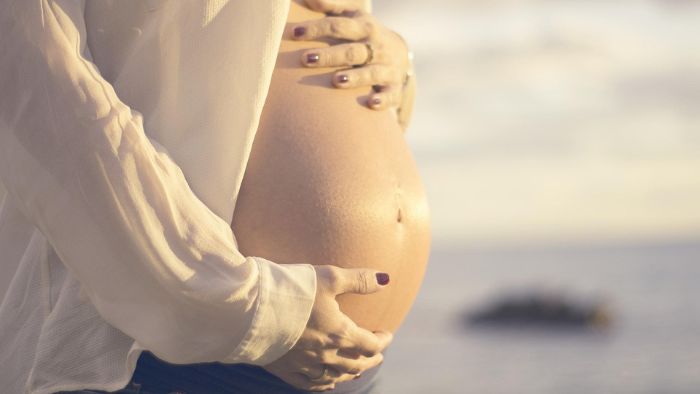 pregnant white woman with her hands on her bare pregnancy bump as she uses the glove anaesthesia hypnobirthing technique with a relaxing beach background behind her