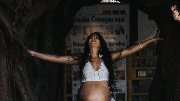 brown pregnant woman with long black hair and wearing a white cropped top lifts her arms our and looks up as she asks how the glove relaxation technique helps birth