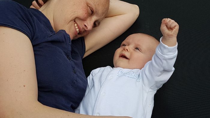 newborn baby rising his right fist as if in celebration with his mum smiling and happy next to him having had a pain free birth using glove anaesthesia in hypnobirthing